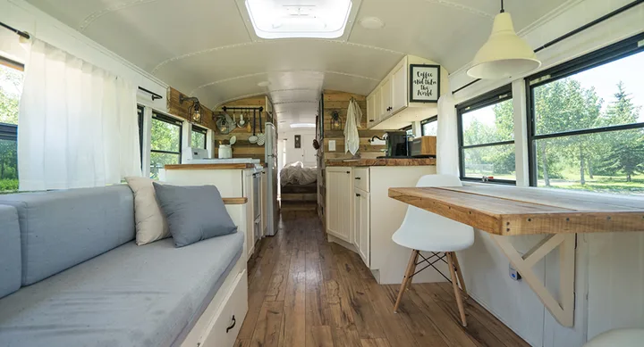 Interior of a converted bus features white and wood decor with a bench, table, and kitchen area under natural light.