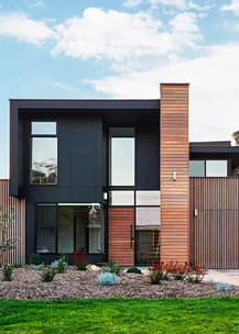 Modern two-story house with wooden and black panel facade, large windows, and landscaped front garden under a blue sky.