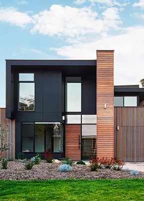 Modern two-story house with wooden and black panel facade, large windows, and landscaped front garden under a blue sky.