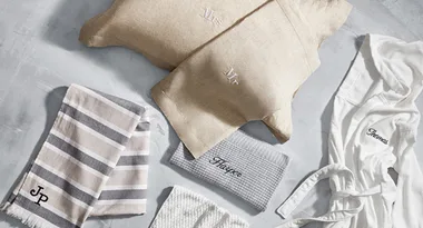 Personalized towels, robes, and pillows with names: "Mr," "Mrs," "Thomas," "Harper," and initials "JP" on a gray surface.