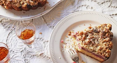 Slice of rhubarb crumble cake on a white plate with sherry glasses on a lace tablecloth.