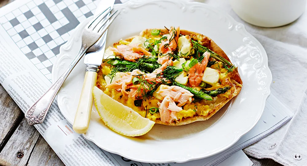 Open-faced sandwich with smoked salmon, asparagus, herbs, and cheese on a white plate with lemon wedge and vintage fork.