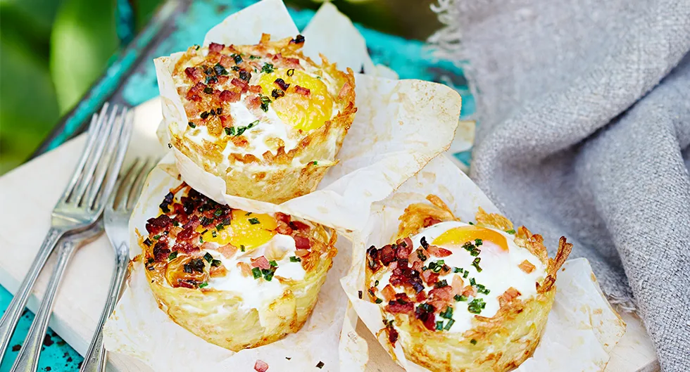 Muffin-shaped hash browns with eggs, bacon, chives on a wooden board with two forks, set outdoors.