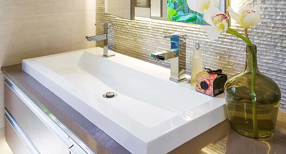 Modern bathroom sink with dual faucets, decorative vase, and a textured wall tile backdrop.