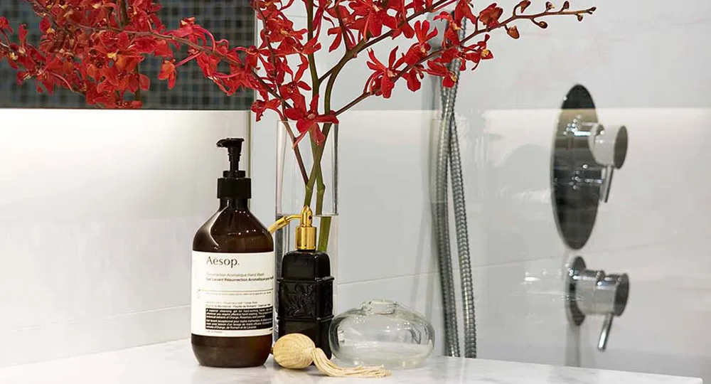 Bottle of Aesop soap, red flowers in vase, and bath decor on bathroom countertop.