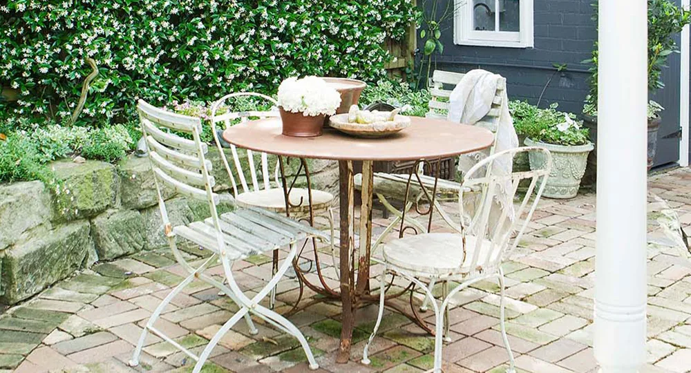 Rustic outdoor patio with round table, mismatched chairs, potted flowers, and lush greenery on a stone paving.
