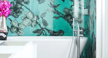 Modern bathroom with a white tub, floral mosaic turquoise tiles, chrome faucet, and pink flowers in a vase.