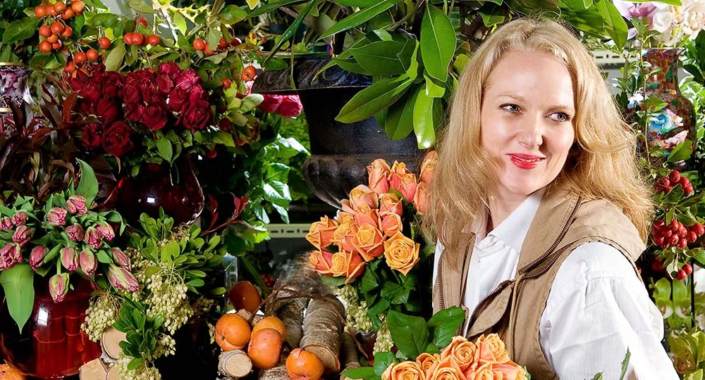 Person arranging flowers in a colorful, lush floral shop setting, surrounded by various vibrant flowers and plants.