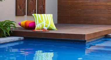 Deck with colorful cushions beside a blue pool, adorned with wall art and a glass of wine on a tray.