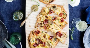 Flatbread with prosciutto, cheese, and herbs on a wooden board, beside a pizza cutter and green bottle, on a blue tablecloth.