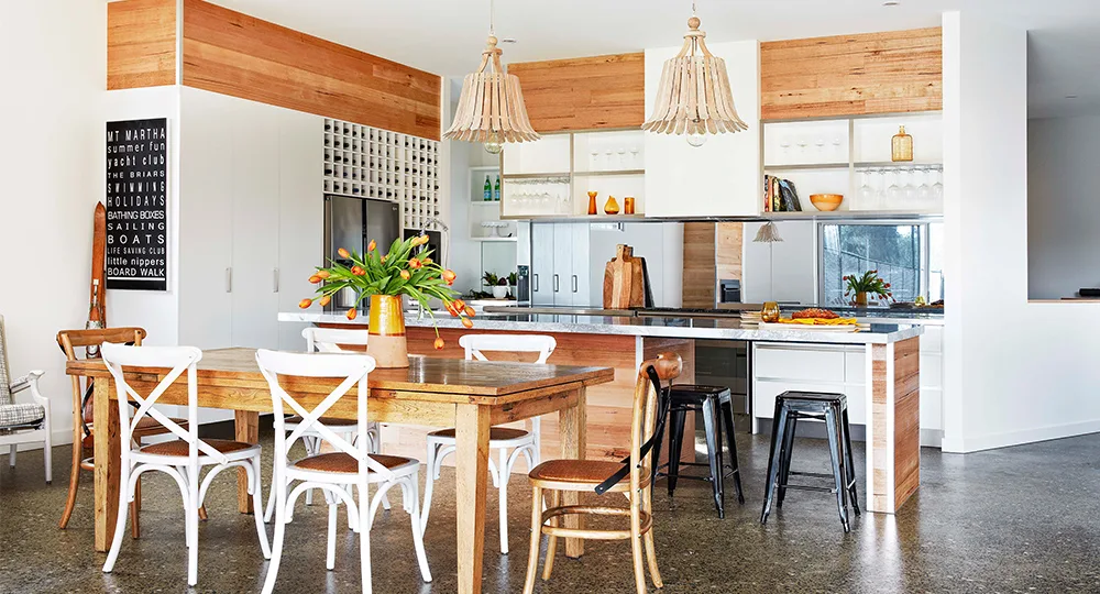 Modern kitchen with wooden dining table, mismatched chairs, pendant lights, and decorative plants.