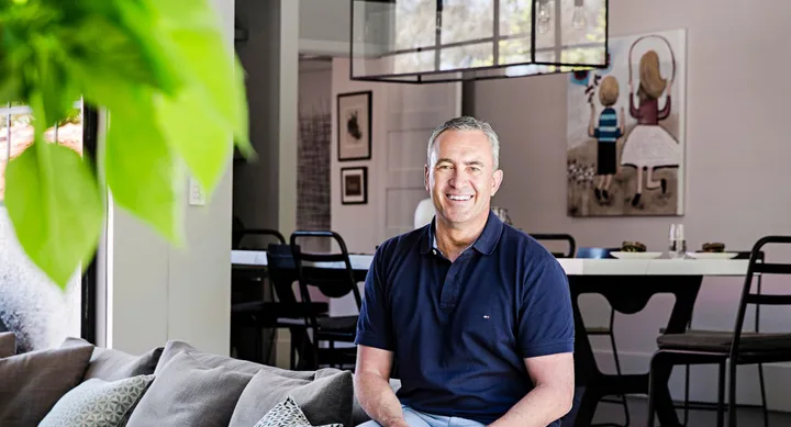 A man smiling, seated on a comfortable couch in a modern, well-lit living room with art on the walls.