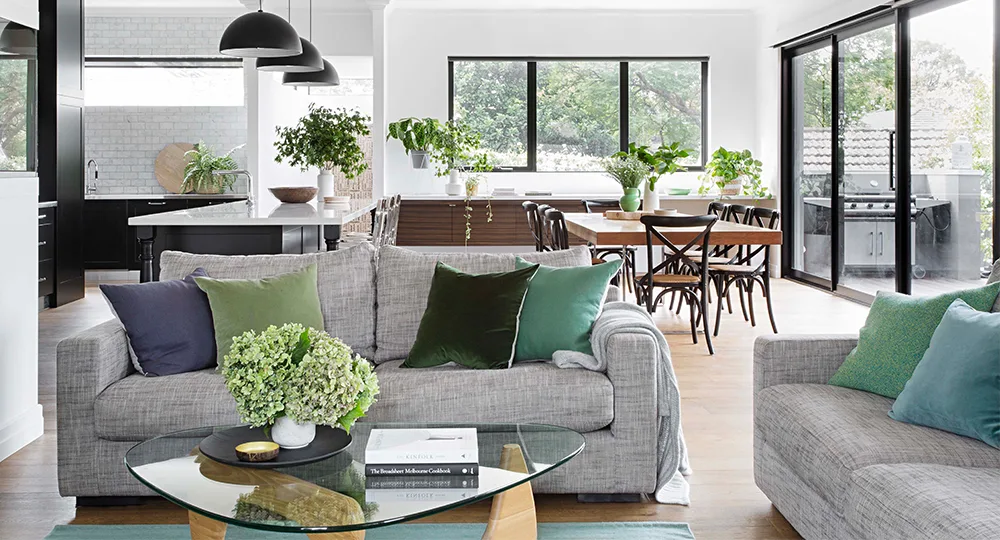 Modern open-plan living area with gray sofas, green cushions, glass coffee table, dining table, and large windows.