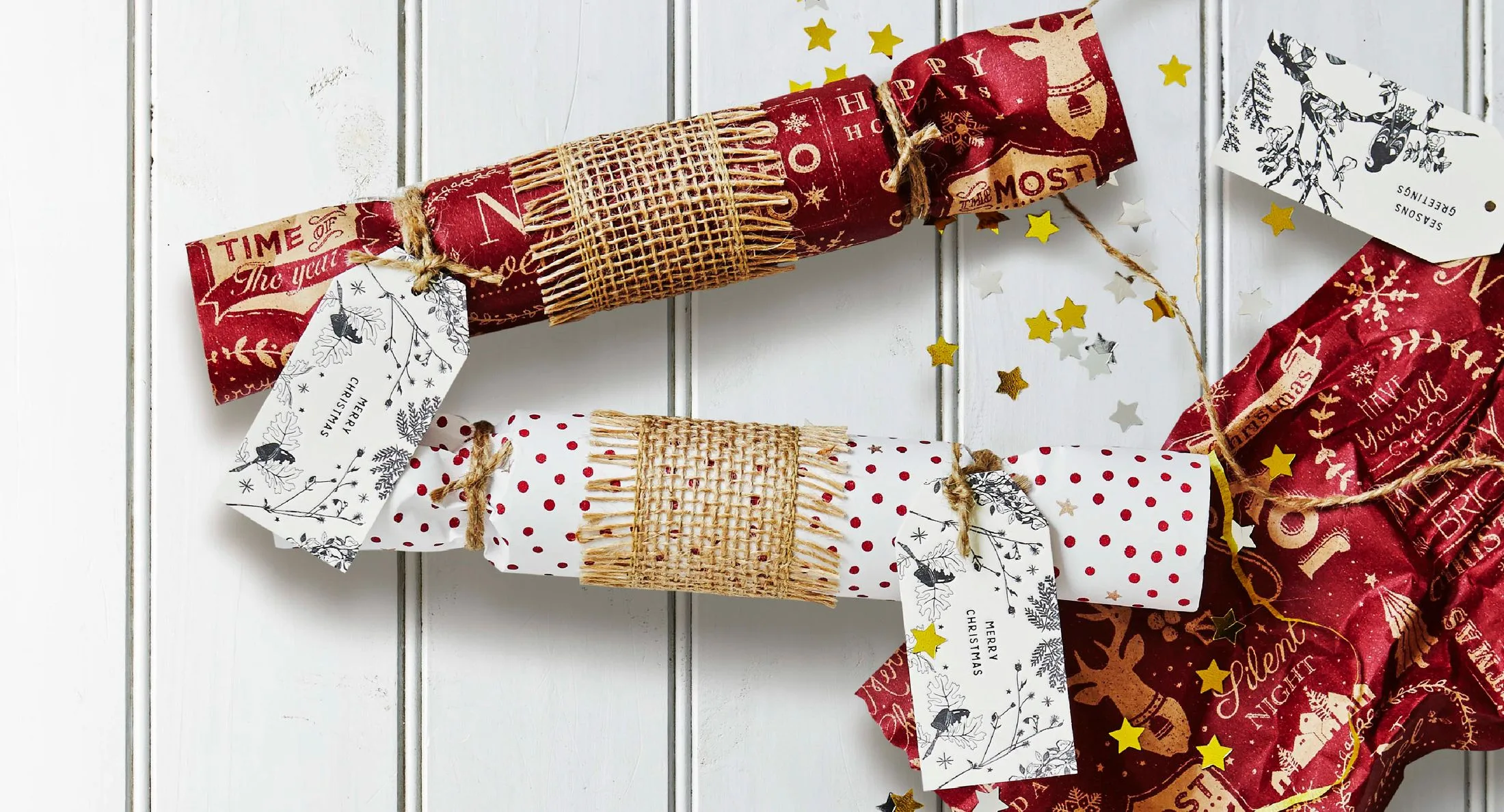 Christmas crackers wrapped in festive paper with tags, placed on a white surface with scattered star confetti.