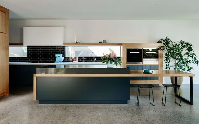 Modern kitchen with black and wood cabinetry, island, blue mixer, plants, and minimal decor.