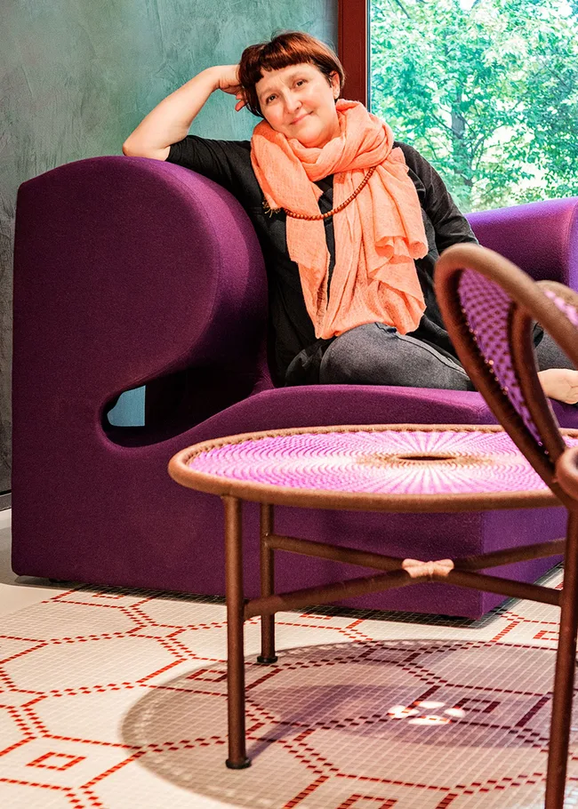 A woman relaxes on a purple chair, wearing a coral scarf, next to a woven table with a geometric rug beneath.