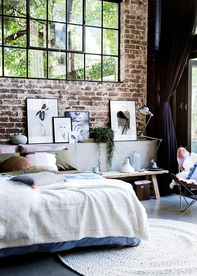 Loft bedroom with a brick wall, large window, cozy bed, art frames, and plants, creating a serene, modern atmosphere.