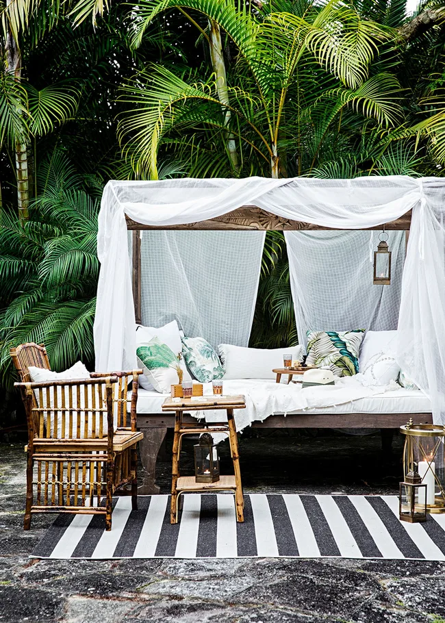 Outdoor canopy bed with white netting, surrounded by lush green plants, wooden chairs, and lanterns on a striped rug.