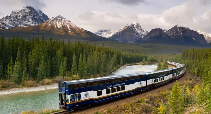 Rocky Mountaineer train traveling through forest with snow-capped mountains in the background.