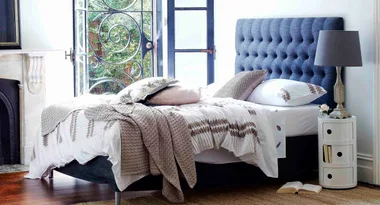 A cozy bedroom with a blue headboard, patterned bedding, and an open window, complete with a bedside lamp and nightstand.