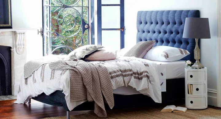 A cozy bedroom with a blue headboard, patterned bedding, and an open window, complete with a bedside lamp and nightstand.