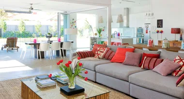 Modern open-plan living room with gray sofa, red cushions, glass table with tulips, and view of outdoor dining area.
