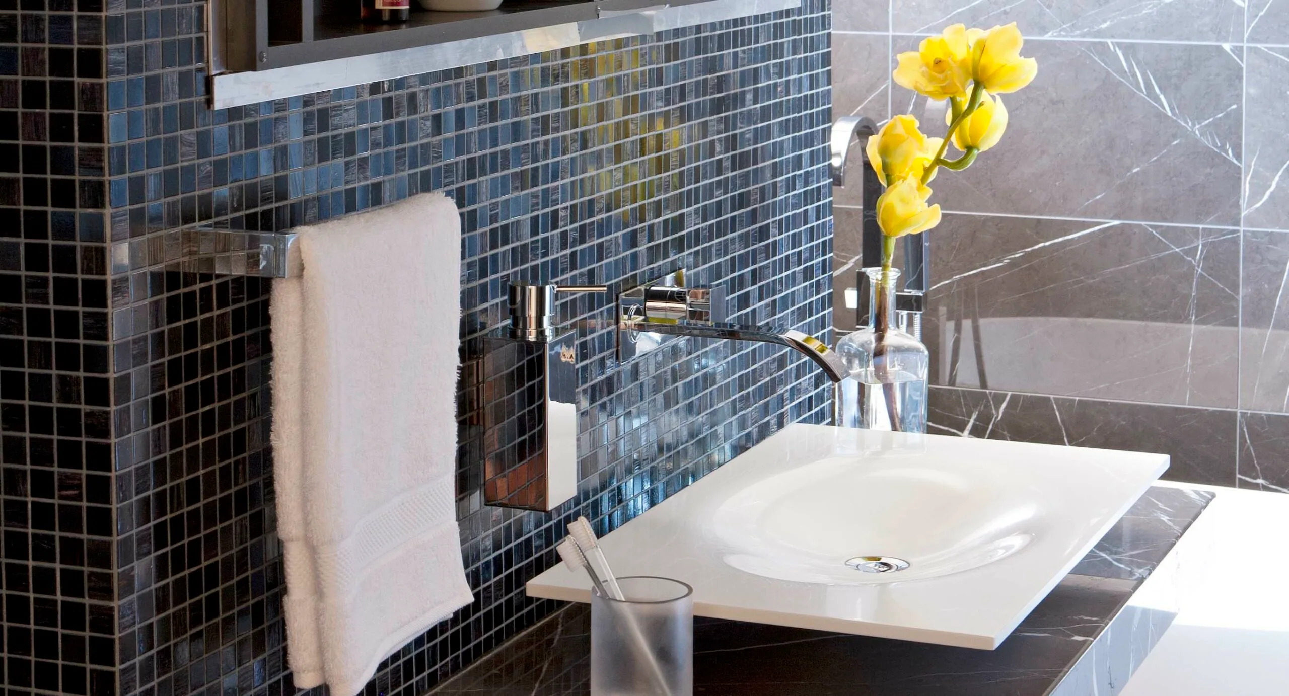 Modern bathroom sink with chrome fixtures, black tiled wall, yellow flowers in a vase, and white towel.