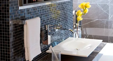 Modern bathroom sink with chrome fixtures, black tiled wall, yellow flowers in a vase, and white towel.