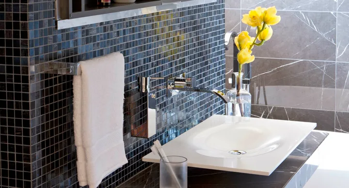 Modern bathroom sink with chrome fixtures, black tiled wall, yellow flowers in a vase, and white towel.