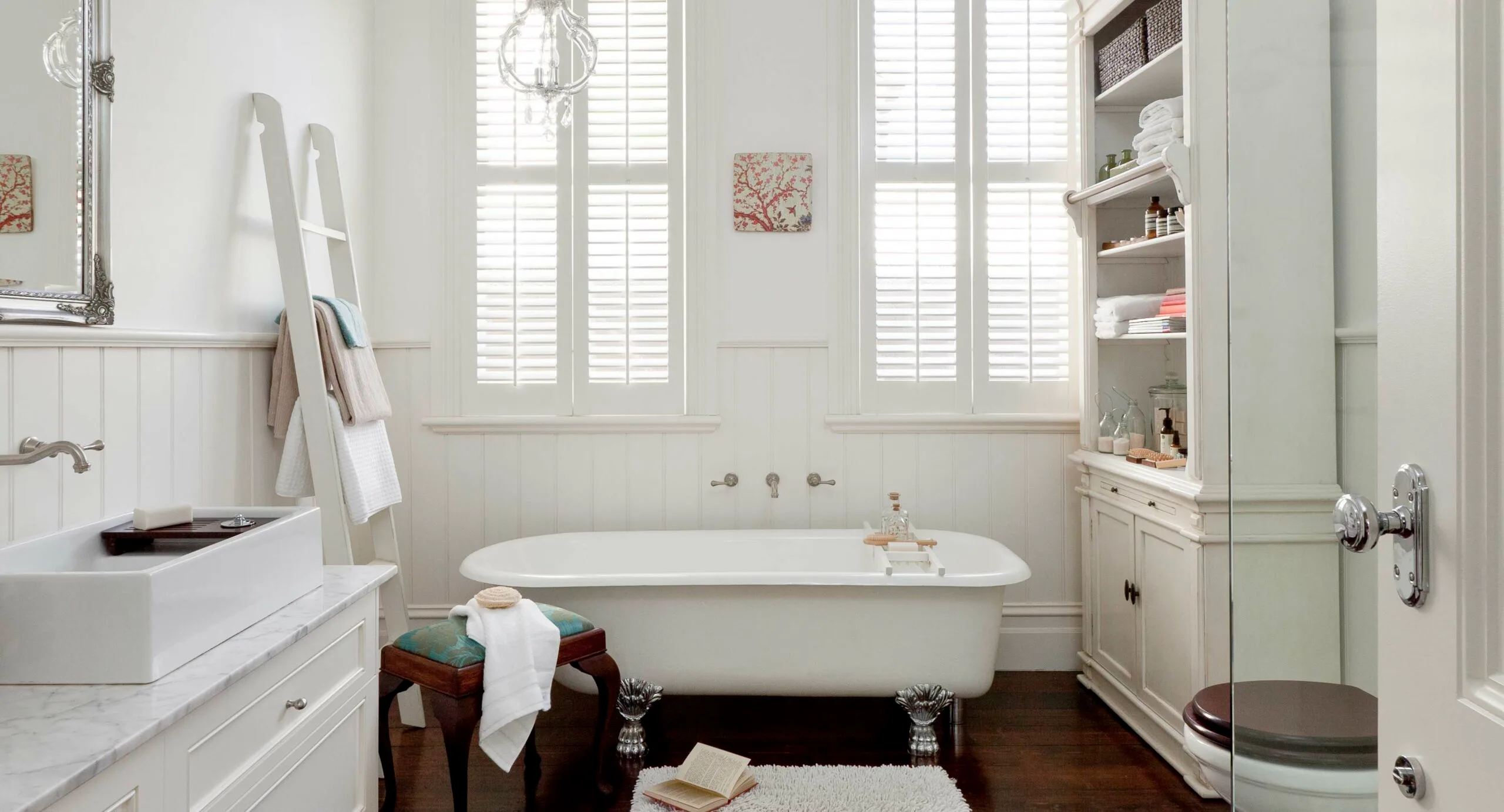Elegant bathroom with a clawfoot tub, white walls, open book, towel ladder, and glass shelving unit by window shutters.