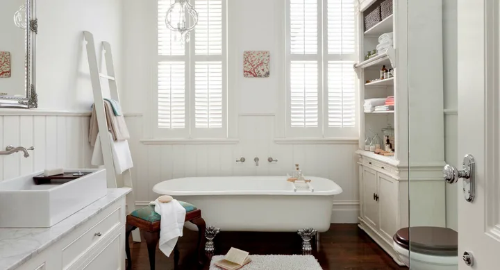 Elegant bathroom with a clawfoot tub, white walls, open book, towel ladder, and glass shelving unit by window shutters.
