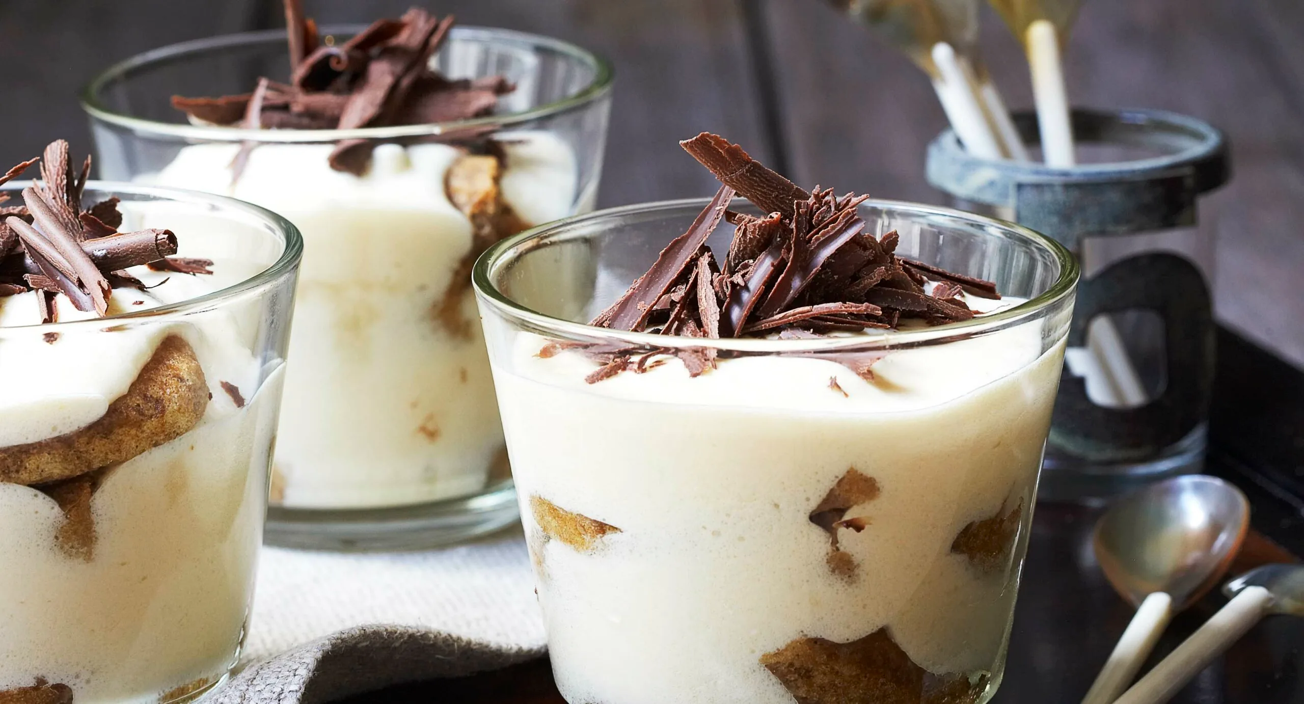 Tiramisu in glass cups topped with chocolate shavings and served with spoons in the background.
