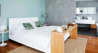 Bedroom with a white bed, tropical pillows, wooden bench, and attached bathroom with double sinks and large mirror.