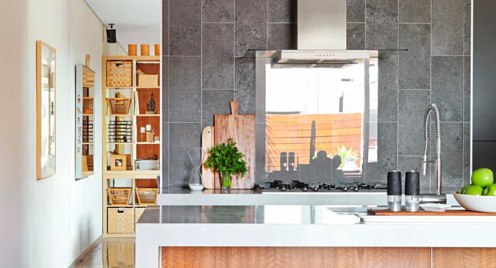 Modern kitchen with gray tiles, a stainless steel stove, chopping boards, potted herbs, apples, and decorative shelving.