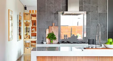 Modern kitchen with gray tiles, a stainless steel stove, chopping boards, potted herbs, apples, and decorative shelving.