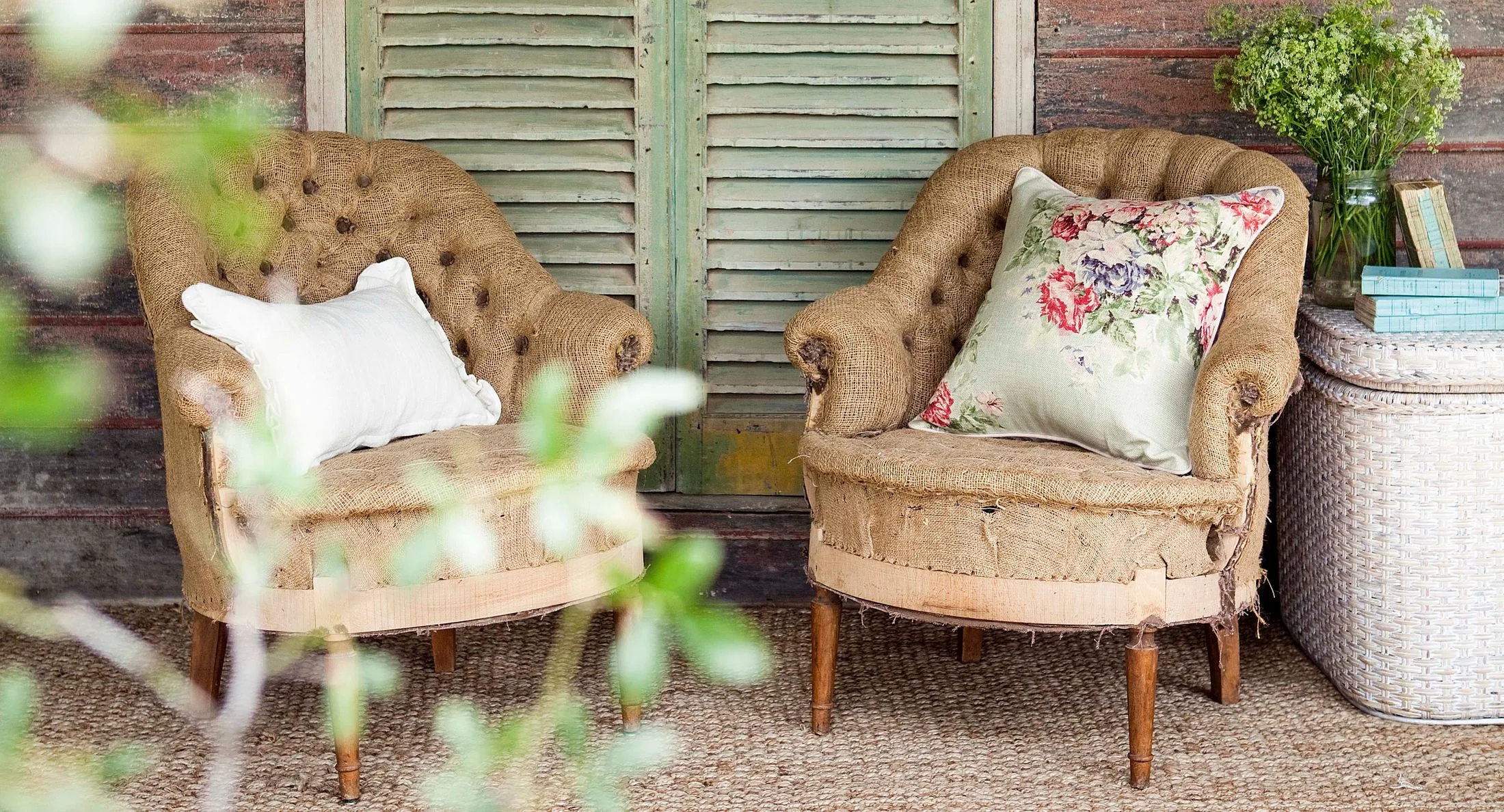 Two rustic armchairs with floral cushions on a porch, framed by greenery, next to a wicker table with books and flowers.