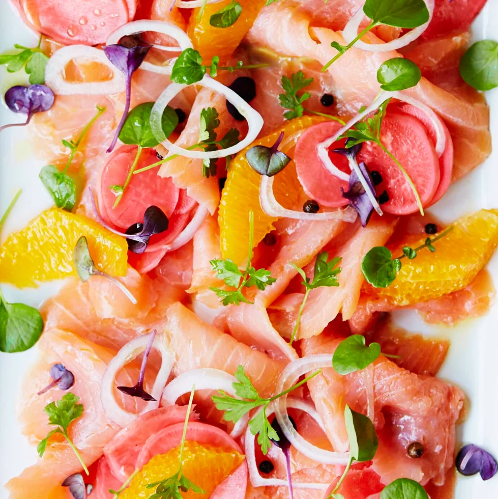 Plate of cured salmon with citrus, radish, microgreens, and onion rings, next to a stack of small forks on a linen cloth.
