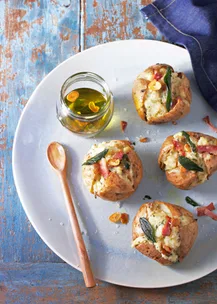 Baked potatoes with bacon, cheese, and sage on a plate, served with a jar of olive oil and a wooden spoon.