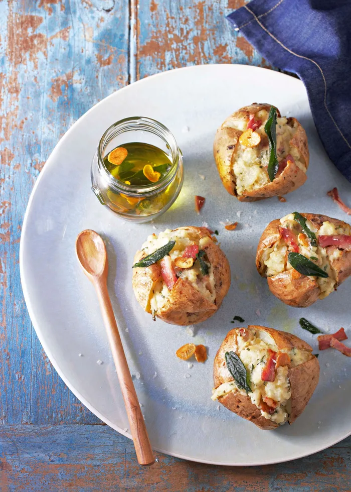 Baked potatoes with bacon, cheese, and sage on a plate, served with a jar of olive oil and a wooden spoon.