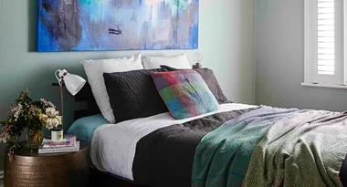 Cozy bedroom with colorful pillows, abstract art, and a side table with a lamp, flowers, and books.