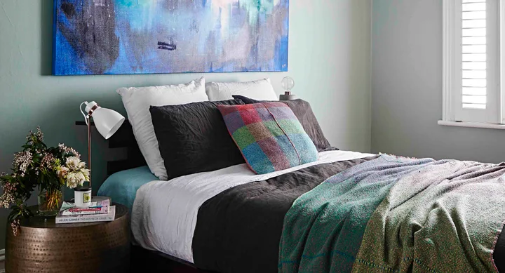 Cozy bedroom with colorful pillows, abstract art, and a side table with a lamp, flowers, and books.