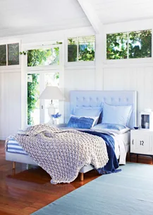 Cozy bedroom with a light blue theme, large windows, plush bedding, and a chunky knit blanket draped on the bed.