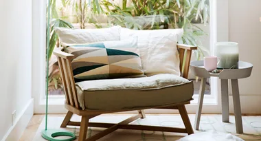 Cozy armchair with geometric pillow and side table with mug and candle, set by a window with green plants outside.