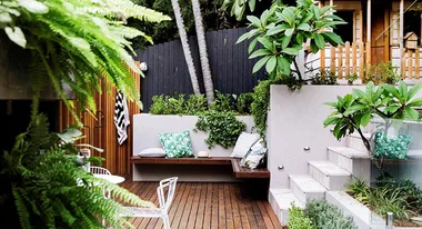 A lush outdoor patio with wooden decking, green plants, a bench with cushions, and a wooden fence in a cozy garden setting.
