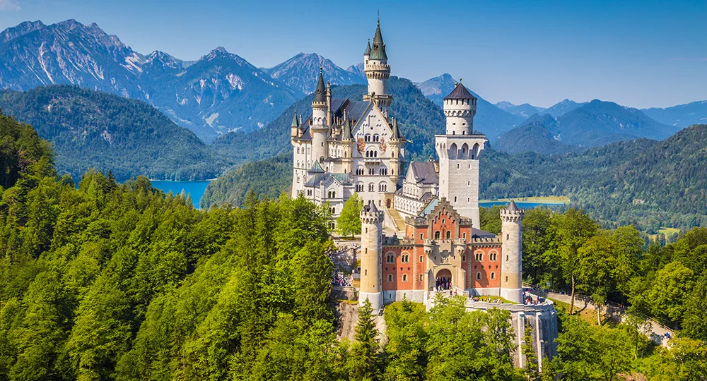 Neuschwanstein Castle surrounded by lush greenery and mountains under a clear blue sky in Bavaria, Germany.