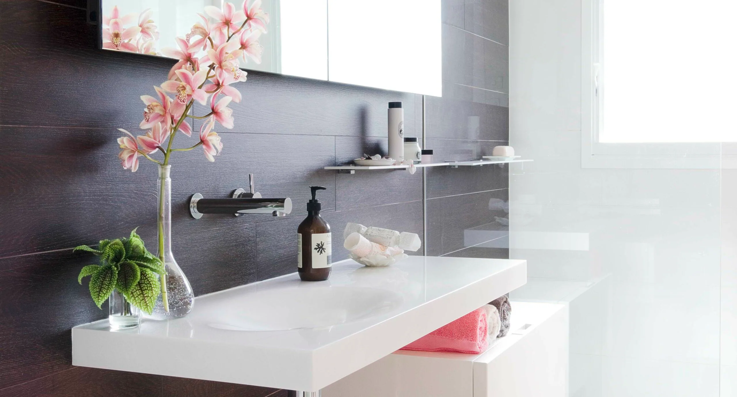 Modern bathroom with white sink, pink orchids, soap dispenser, and neatly folded towels under a bright window.