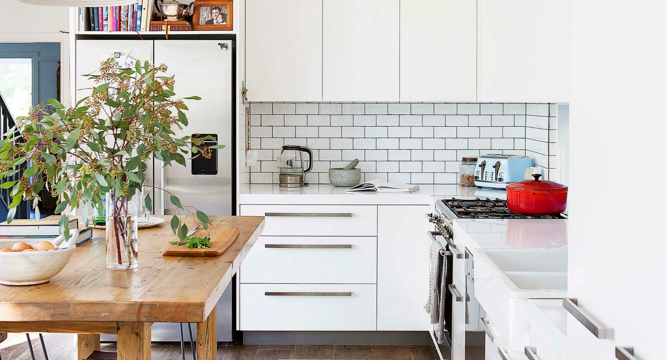 Modern kitchen with wooden table, vase with greenery, white cabinets, and stainless steel fridge.