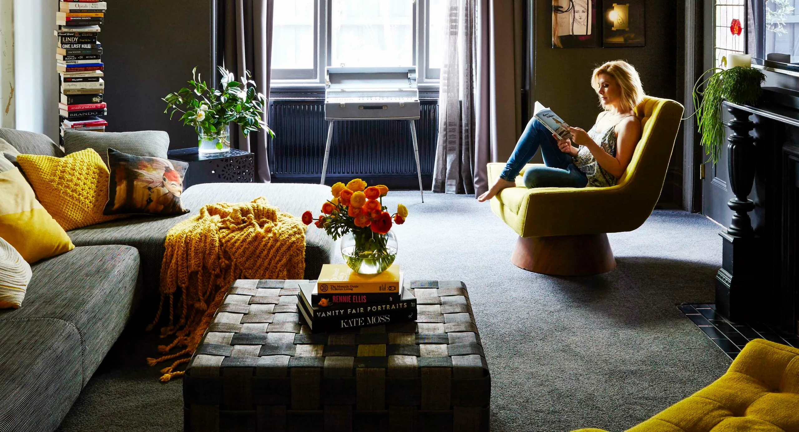 Woman reading on a yellow chair in cozy living room with books, plants, and colorful decor accents.