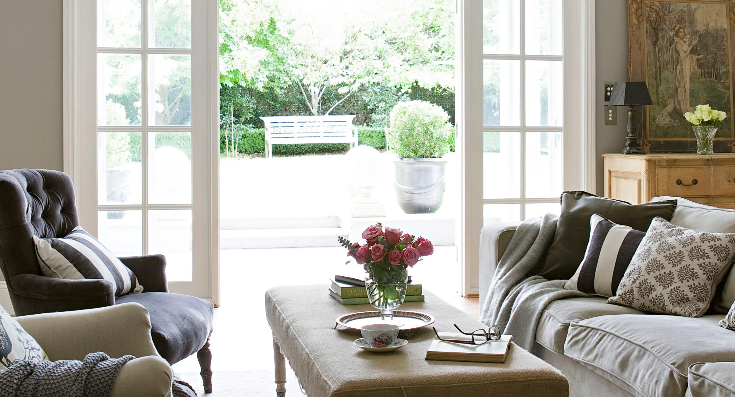 Living room with cozy seating, cushions, a coffee table with flowers, and large windows opening to a green garden.