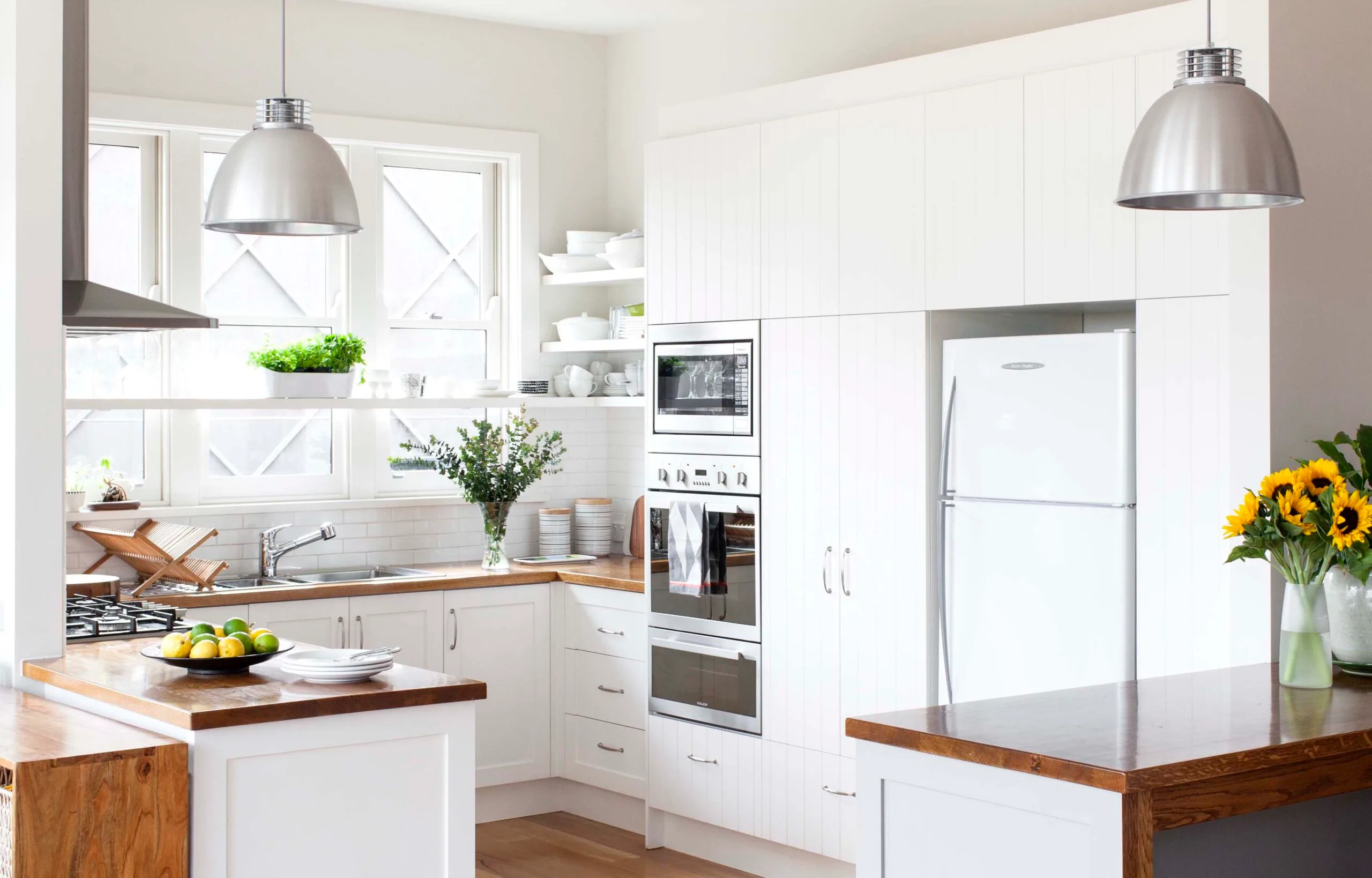 Modern white kitchen with wood countertops, stainless steel appliances, and pendant lights; sunflowers on the island.
